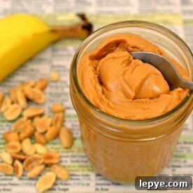 A pint jar of freshly made peanut butter with a spoon in it, next to a banana and whole peanuts.