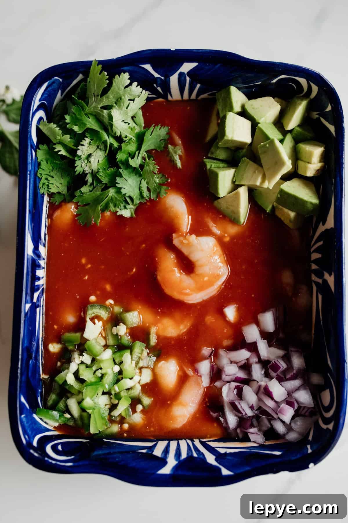 Mexican Shrimp Cocktail Recipe: Zesty Camarones en Coctel 6 All ingredients for Mexican Coctel de Camarones placed in a rectangular blue serving dish, including cooked shrimp, avocado, red onion, cilantro, lime, and a tomato-based sauce, ready to be mixed