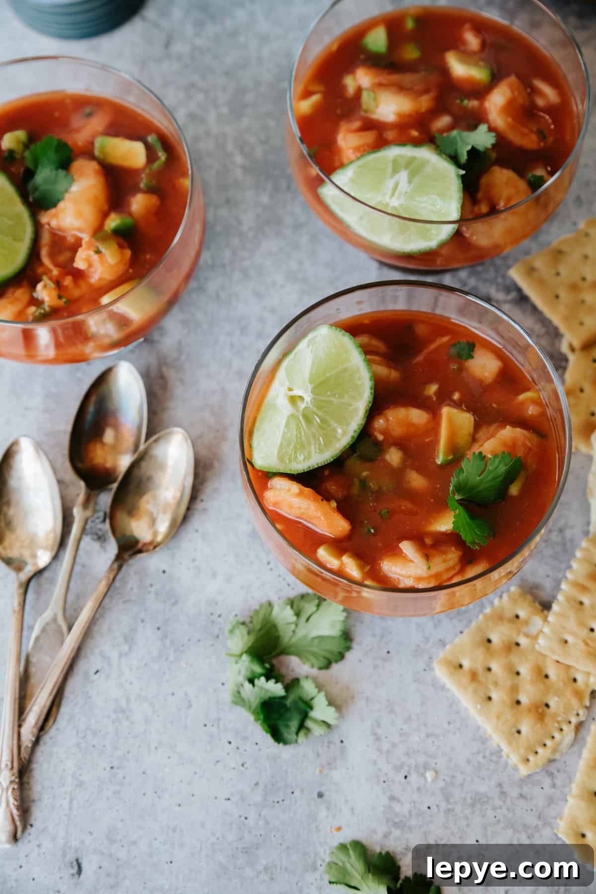 Mexican Shrimp Cocktail Recipe: Zesty Camarones en Coctel 8 Mexican shrimp cocktail (coctel de camarones) served in glass cups with shrimp, avocado, cilantro, and lime in a tomato-based sauce, surrounded by saltine crackers and vintage spoons