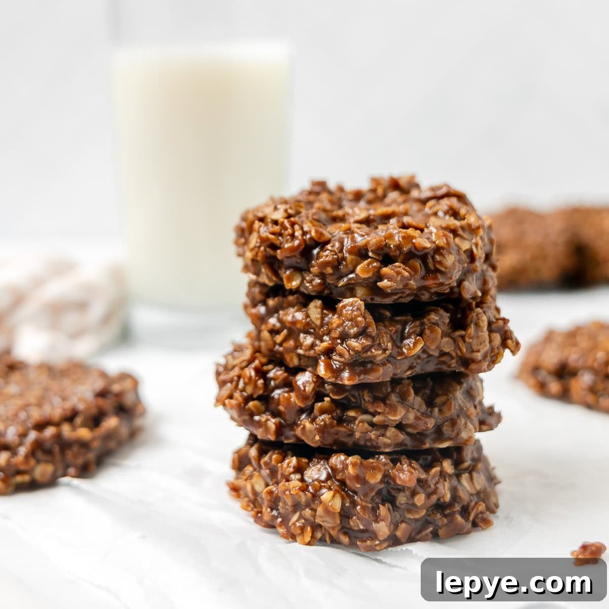A stack of four chocolate oat cookies sits on white parchment paper, with a glass of milk in the background.