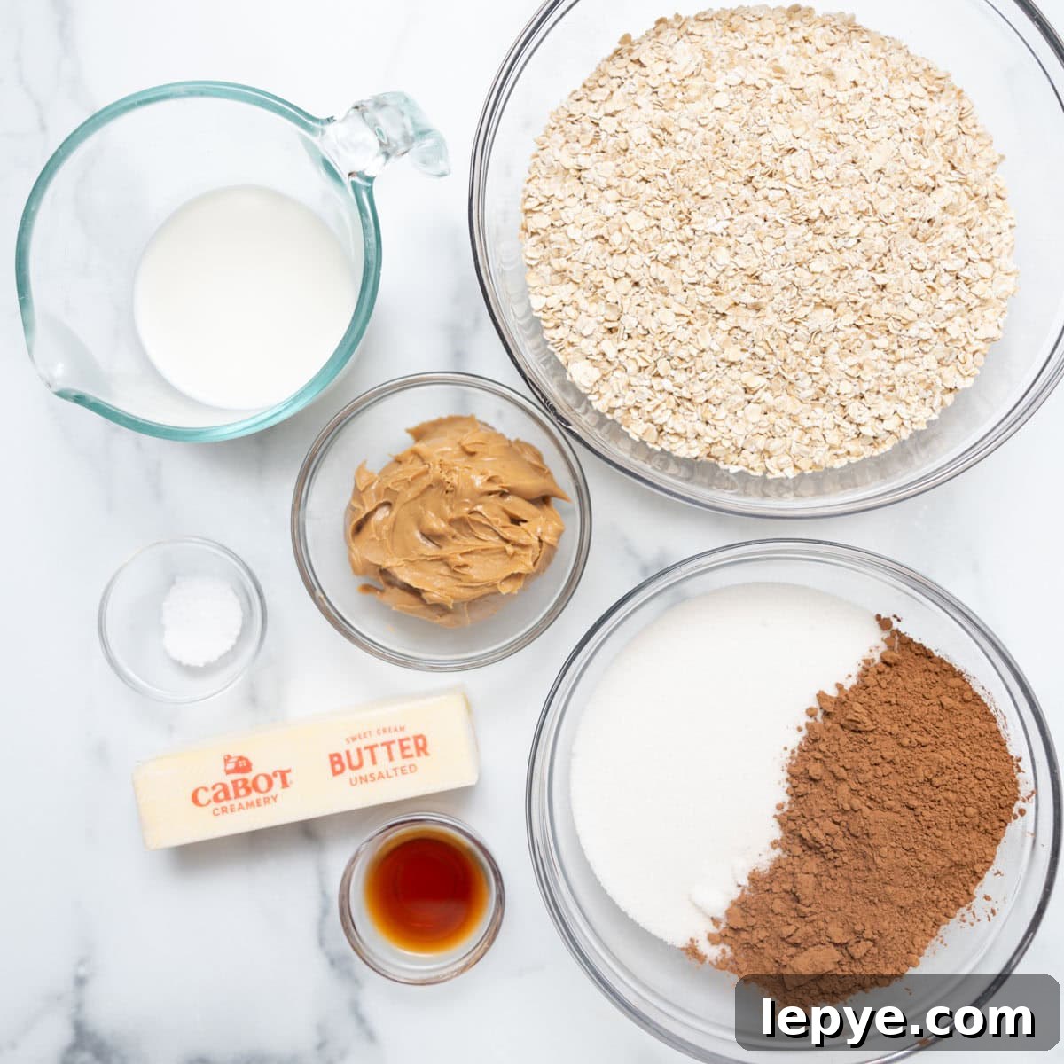 Bowls and containers filled with oats, milk, peanut butter, cocoa powder, sugar, vanilla extract, butter, and salt are arranged on a white marble surface.