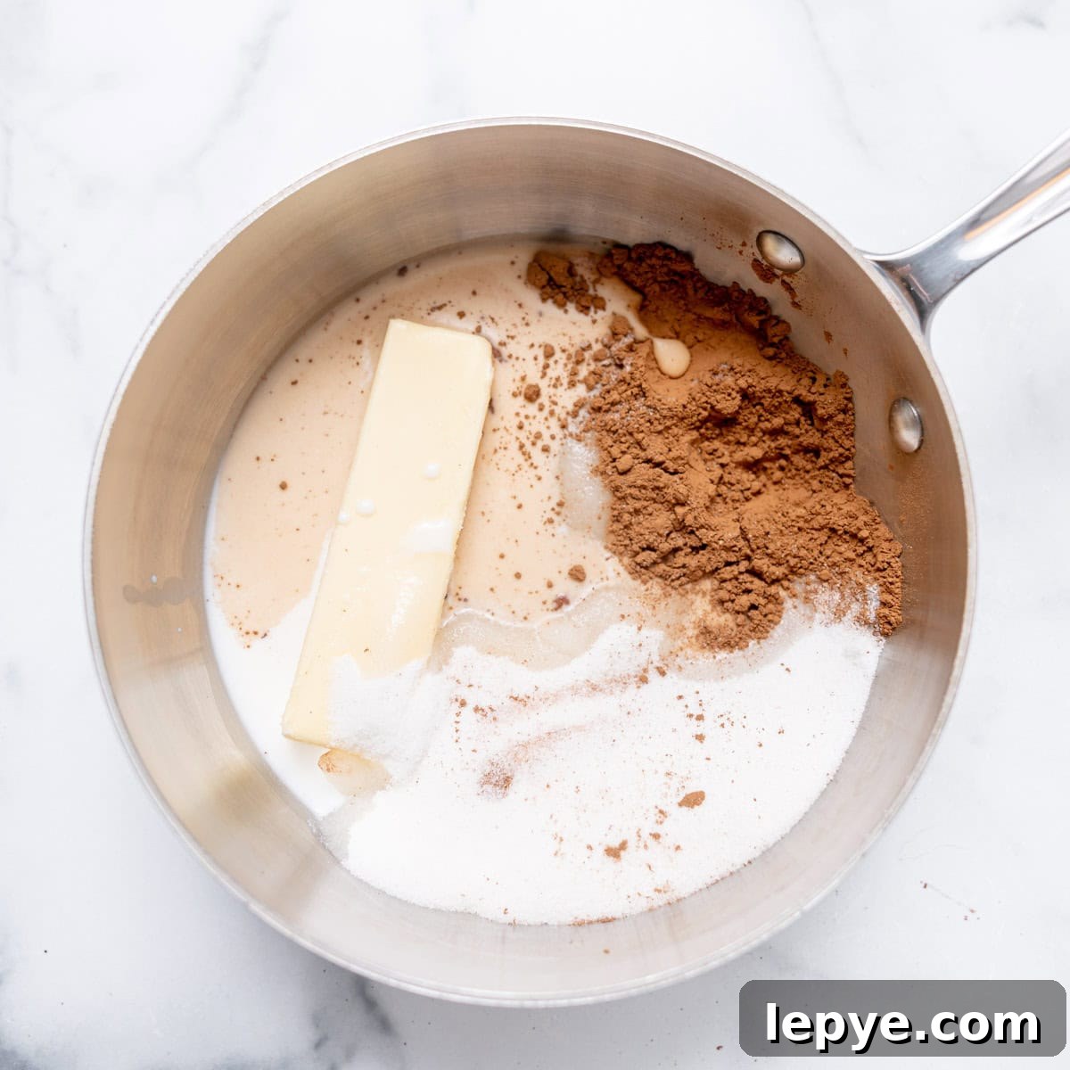A saucepan on a marble surface containing a stick of butter, cocoa powder, sugar, and milk, not yet mixed.