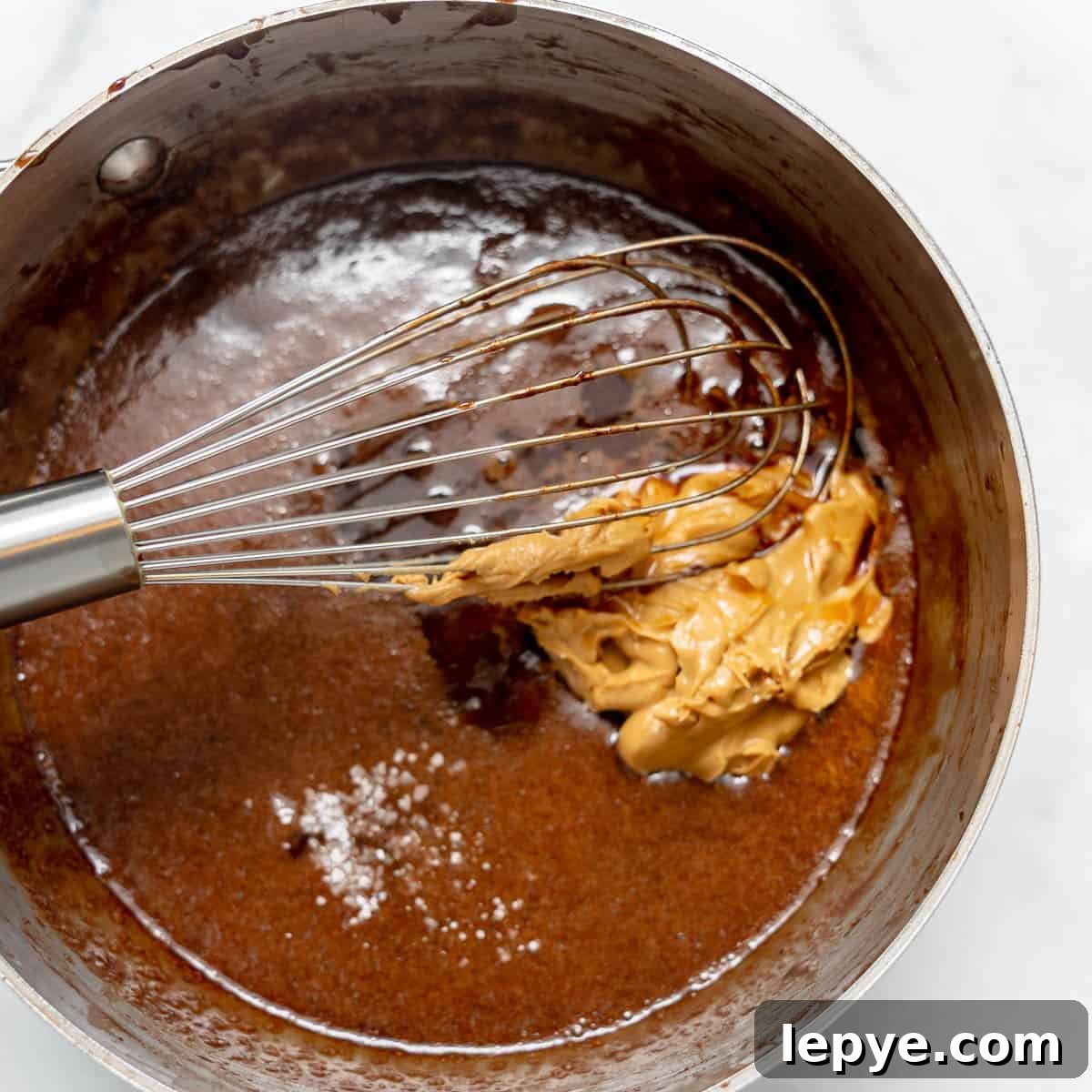 A metal whisk in a saucepan mixing chocolate batter with a dollop of peanut butter and a sprinkle of salt.