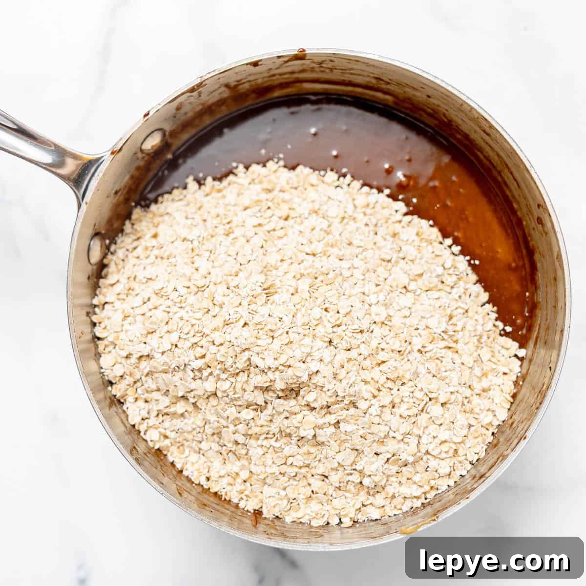 A saucepan filled with chocolate mixture and a layer of dry oats on top, placed on a white marble surface.