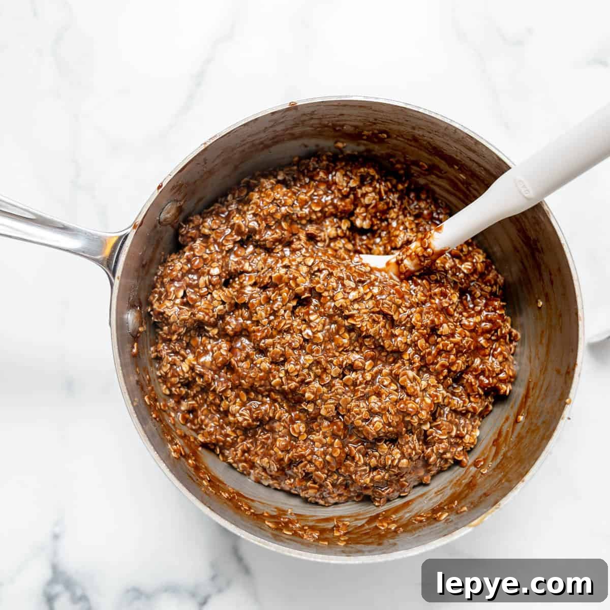 A saucepan filled with a chocolate oat mixture being stirred with a white spatula on a marble surface.