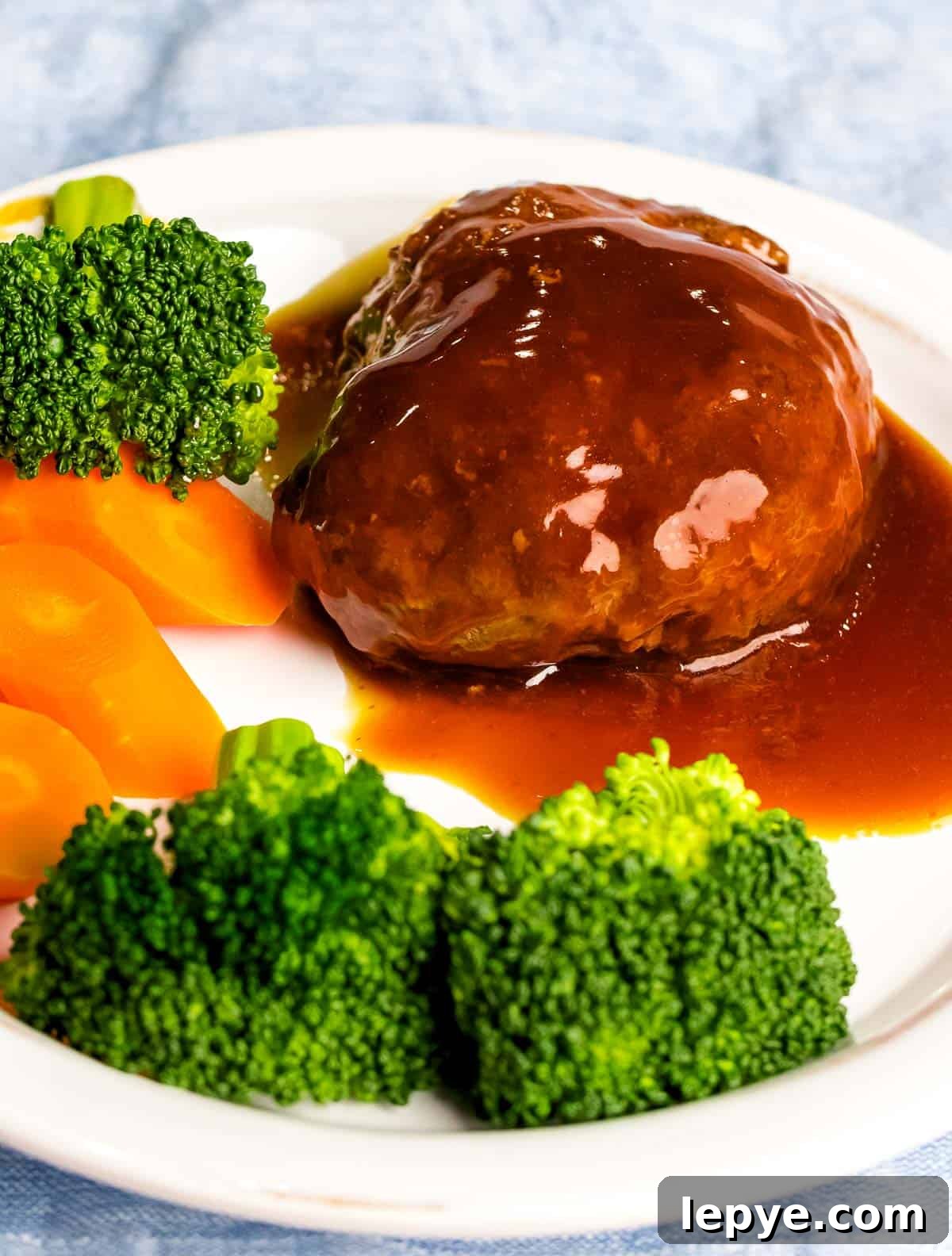 Juicy, tender Japanese hamburger steak (hambāgu ハンバーグ) covered in tangy brown gravy on a white plate next to carrots and broccoli.