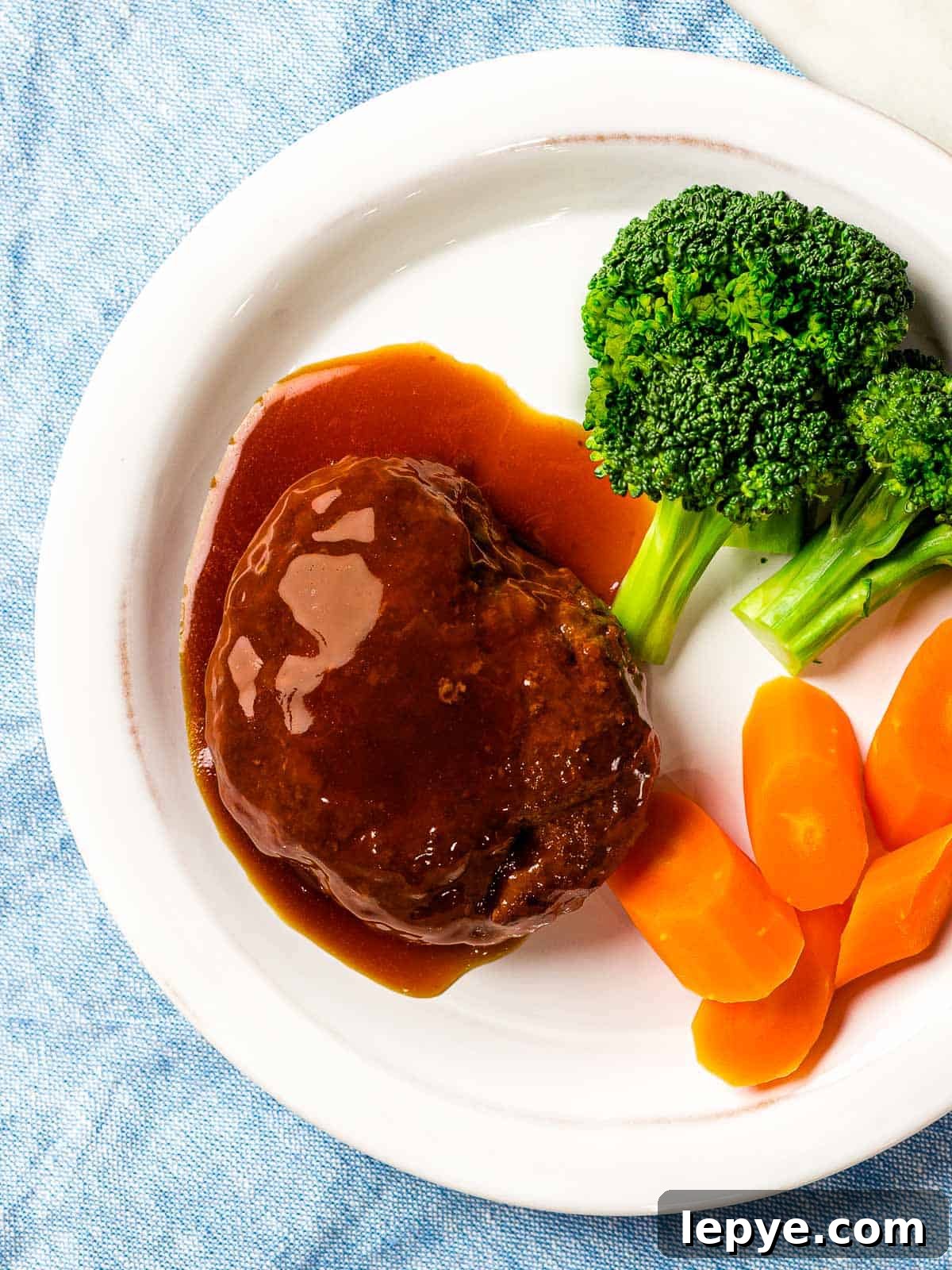 Japanese hamburger steak (hambāgu ハンバーグ) covered in tangy brown gravy on a white plate next to carrots and broccoli.