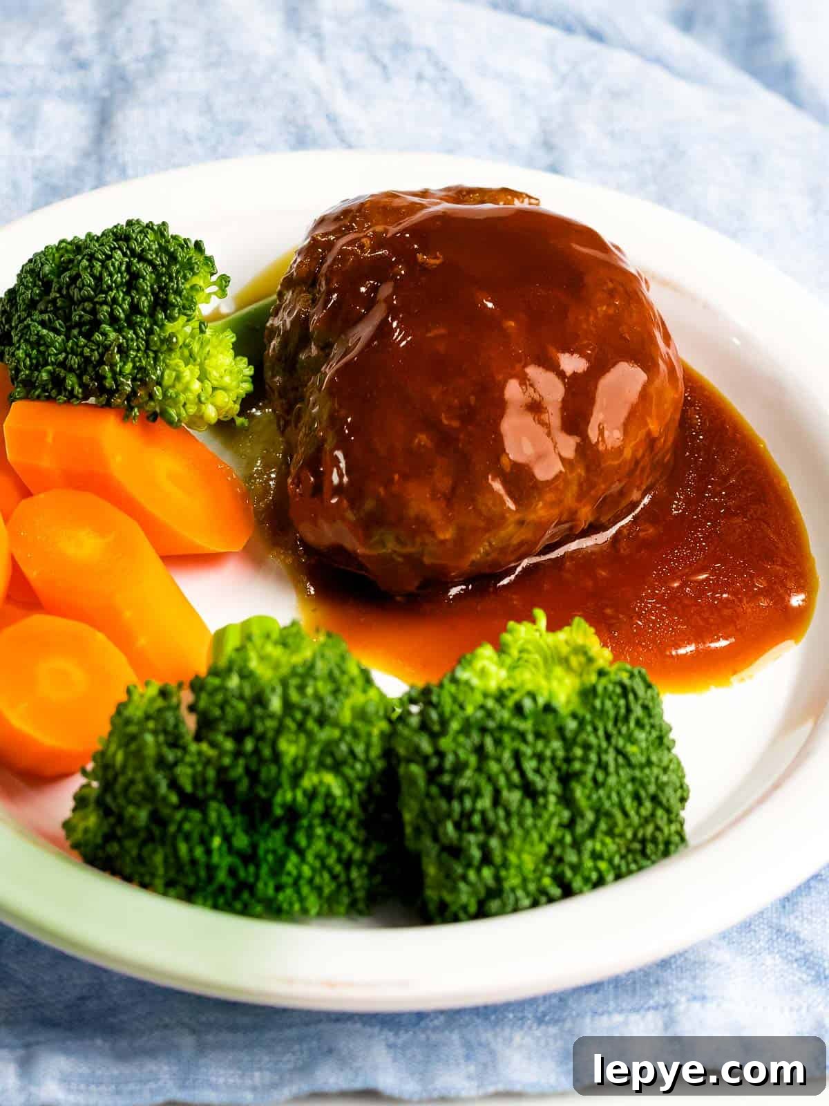 Juicy, tender Japanese hamburger steak (hambāgu ハンバーグ) covered in tangy gravy on a white plate next to carrots and broccoli.