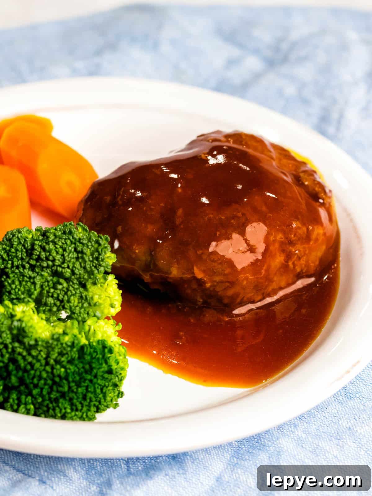 Juicy Japanese hamburger steak (hambāgu ハンバーグ) covered in sauce on a white plate next to carrots and broccoli.