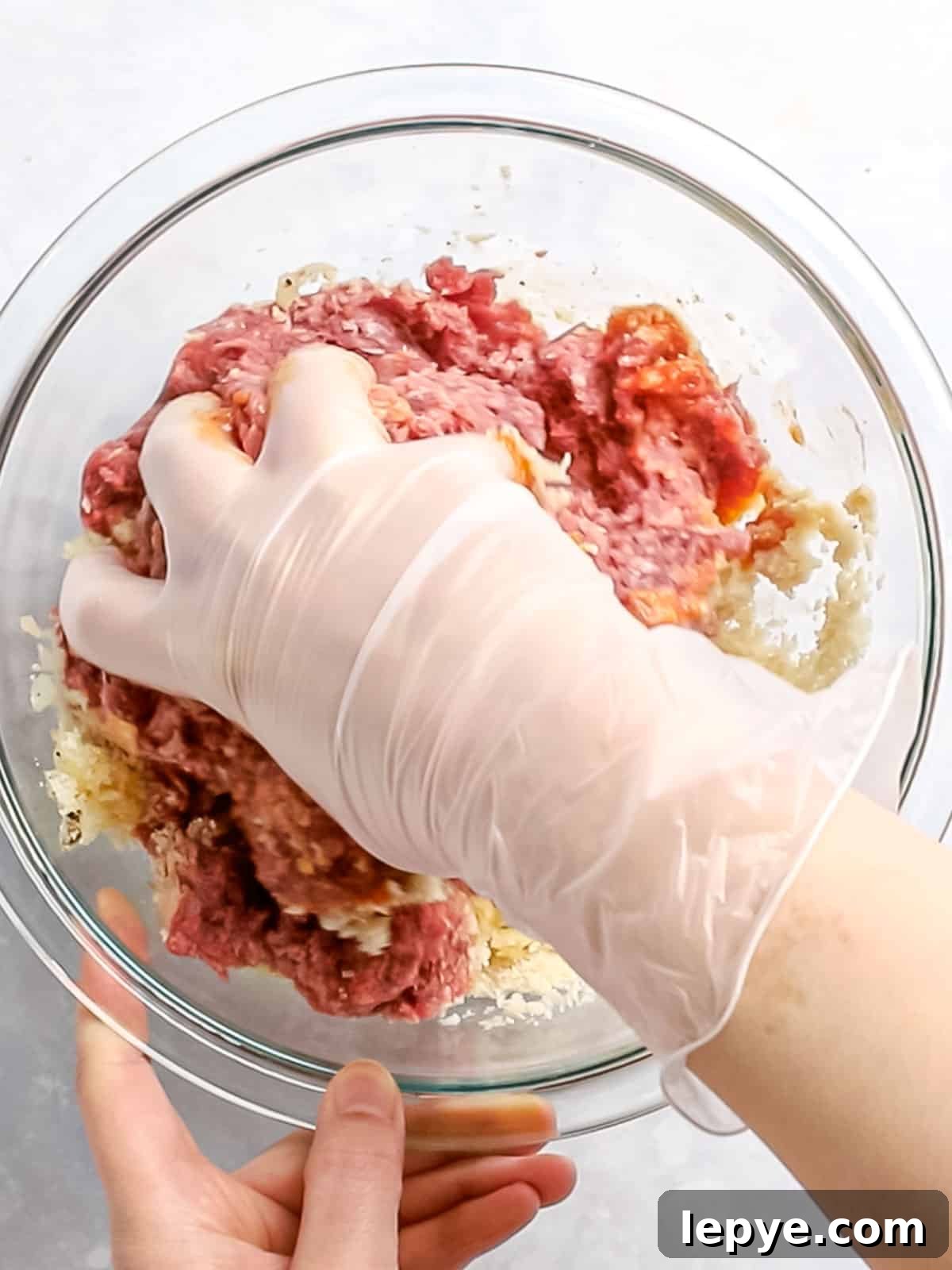 Hamburger steak ingredients in a glass bowl being mixed together with a gloved hand.