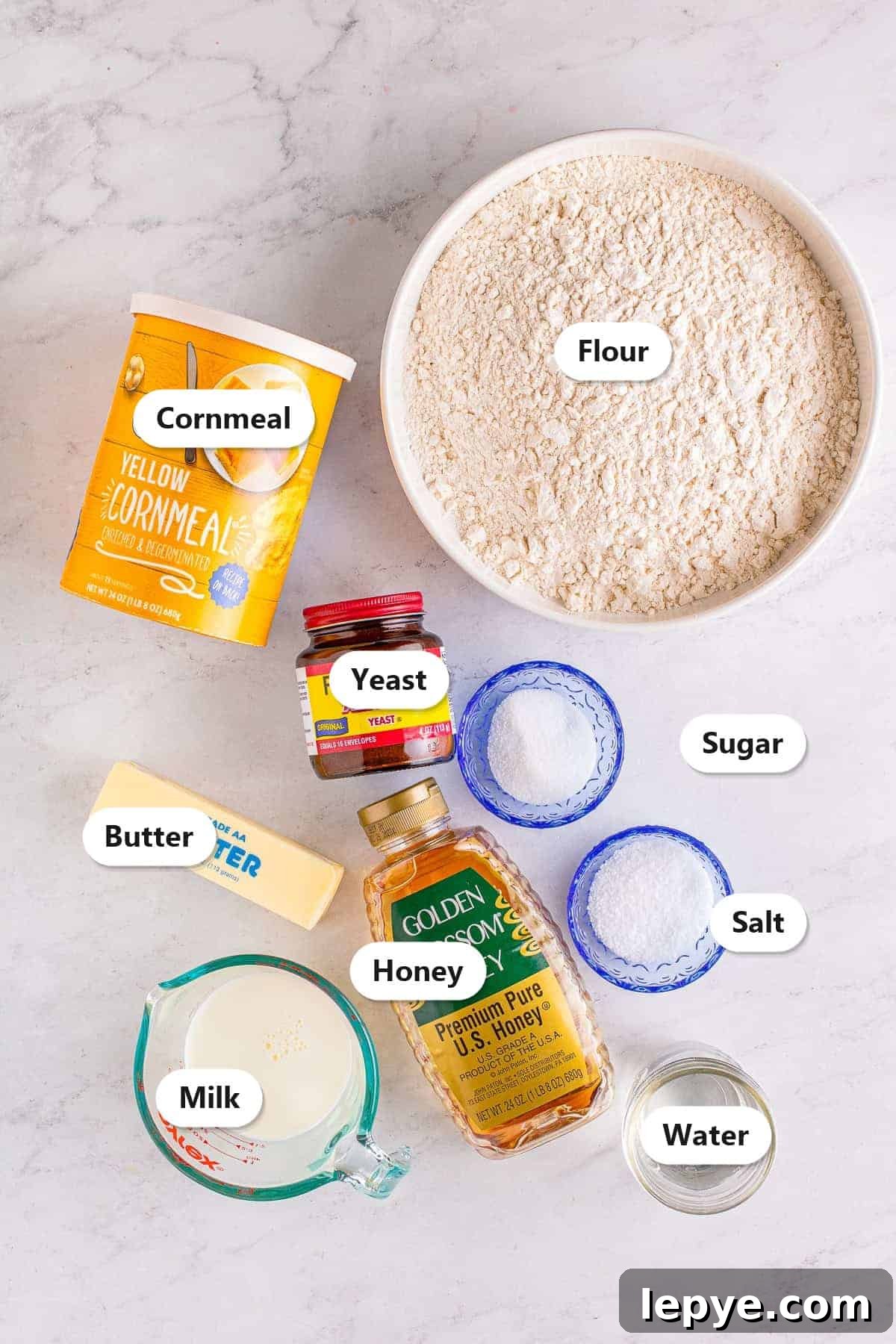 Labeled ingredients to make English muffins including flour, yeast, sugar, salt, milk, water, honey, butter, and cornmeal.