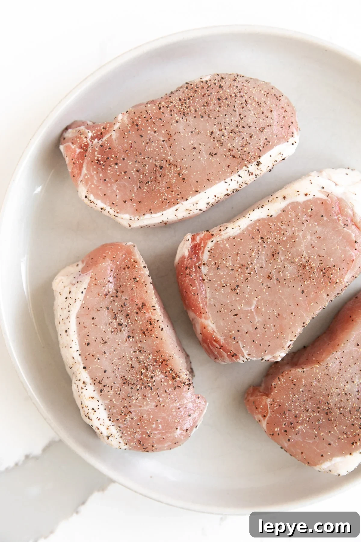Image of four thick-cut pork chops on a plate sprinkled with salt and pepper.